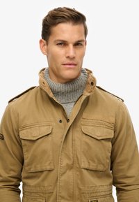 Beige katoenen jack met vier voorknopzakken, een kraag gevoerd met corduroy, en daaronder een gestructureerde grijze coltrui.