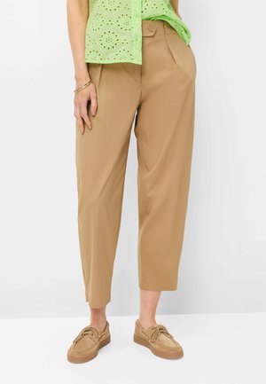 Femme portant un pantalon court beige, des chaussures à lacets marron clair et un haut à manches courtes en broderie anglaise vert clair, debout avec une main dans la poche.