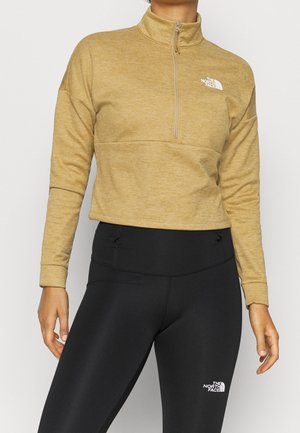 Person iført tan quarter-zip pullover og sorte leggings, begge med The North Face-logo på bryst og lår.