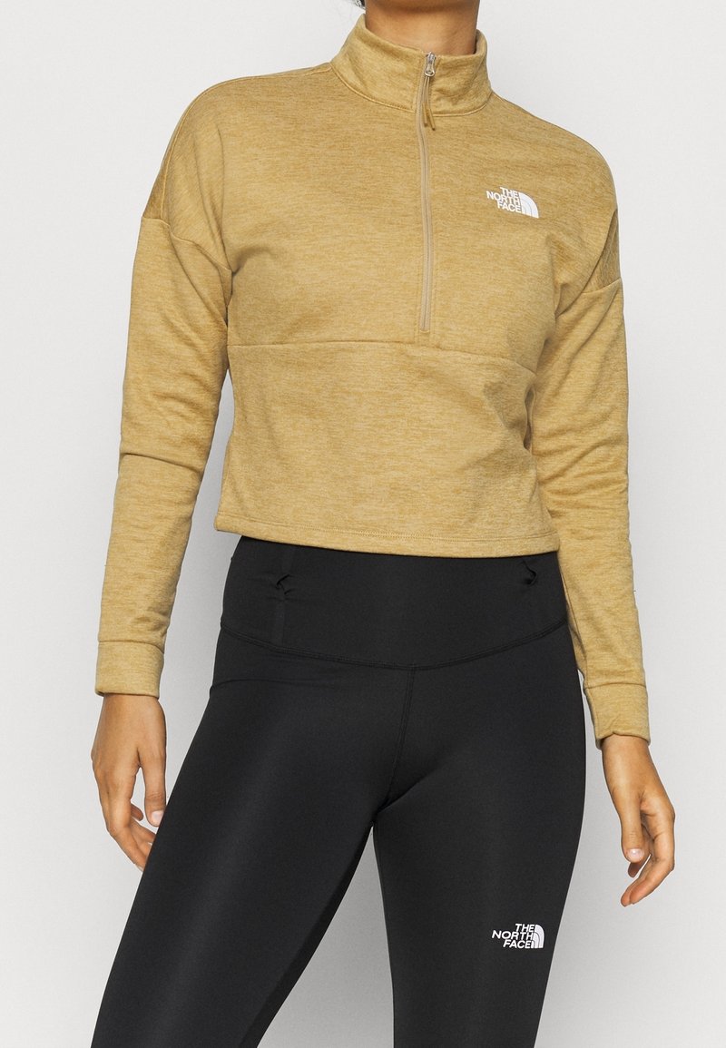 Personne portant un pull beige à fermeture éclair quart et des leggings noirs, tous deux avec le logo The North Face sur la poitrine et la cuisse.
