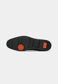 Suela de zapato negra con superficie texturizada, que presenta un acento circular naranja y marca. Diseño estructurado con múltiples patrones de la banda de rodadura.