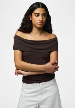Kvinde med langt, krøllet hår har en off-shoulder brun top og hvide bukser på, står med korslagte arme og kigger til siden.