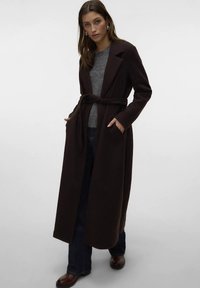 Garš brūns mētelis ar jostu, atloku apkakli un sānu kabatām, valkāts virs pelēka adīta džempera un tumšām džinsām. Gluda auduma tekstūra.