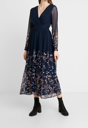 Frau trägt ein marineblaues Midikleid mit langen Ärmeln, floralem Druck auf transparenten Ärmeln und Rock, kombiniert mit schwarzen Stiefeletten.