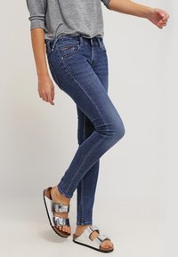 Jeans skinny de denim azul oscuro, con una cintura de altura media y un ligero desvanecimiento. Combinados con sandalias plateadas y negras con dos hebillas.