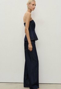 Denimová top bez ramínek s peplum detailem, spárovaný s kalhotami širokého střihu. Tmavě modrý materiál s kontrastním prošíváním a bočními kapsami. Hladká textura.