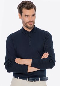 Marineblaues Langarm-Polohemd mit einer Drei-Knopf-Leiste. Weicher Strickstoff, gerippte Bündchen und klassisches Kragen-Design. Trägt einen beigen Gürtel.