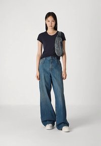 Temno modra majica z širokimi modrimi jeans hlačami. Model drži sivo denim torbo, ki je premeščena čez eno ramo. Beli supergi dopolnjujejo obleko.