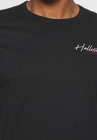 Svart bomullströja med rund halsringning, prydd med vit och röd broderad text som säger "Hollister CALIFORNIA" på bröstet.