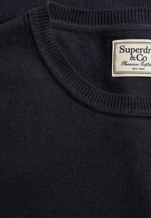 Pull en maille bleu marine foncé avec col rond côtelé et étiquette rectangulaire blanche indiquant "Superdry & Co Premium Outfitters New York."