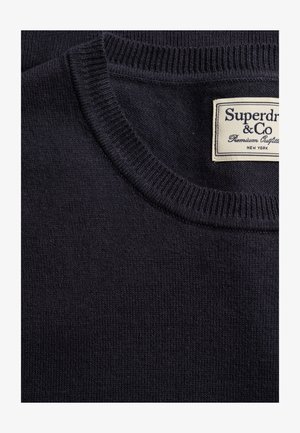 Ciemnogranatowy sweter dziany z prążkowanym okrągłym dekoltem oraz białą prostokątną metką z napisem "Superdry & Co Premium Outfitters New York."