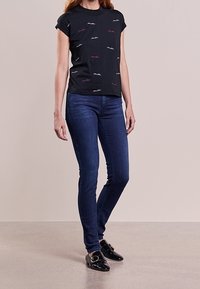 T-shirt nera a maniche corte con linee ricamate colorate, abbinata a jeans skinny blu scuro e mocassini neri con fibbia decorativa.