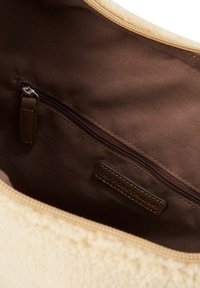 Beige stoffen tas met een bruine binnenkant, voorzien van een ritsvak. Glad materiaal met een gestructureerde rand en een merklabel.
