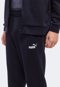 Gros plan d'une personne portant un pantalon de sport Puma noir et une veste noire zippée avec le logo Puma visible sur le pantalon.