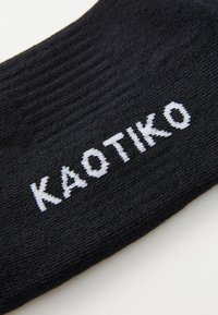 Juodas kojinės, su briaunuota struktūra, su baltu siuvinėtu tekstu "KAOTIKO", ryškiai matomu šone.