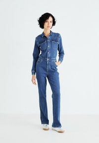 Combinaison en denim à manches longues, boutonnée à l'avant, avec deux poches poitrine, couleur bleue, taille cintrée et coupe droite. Baskets argentées portées.