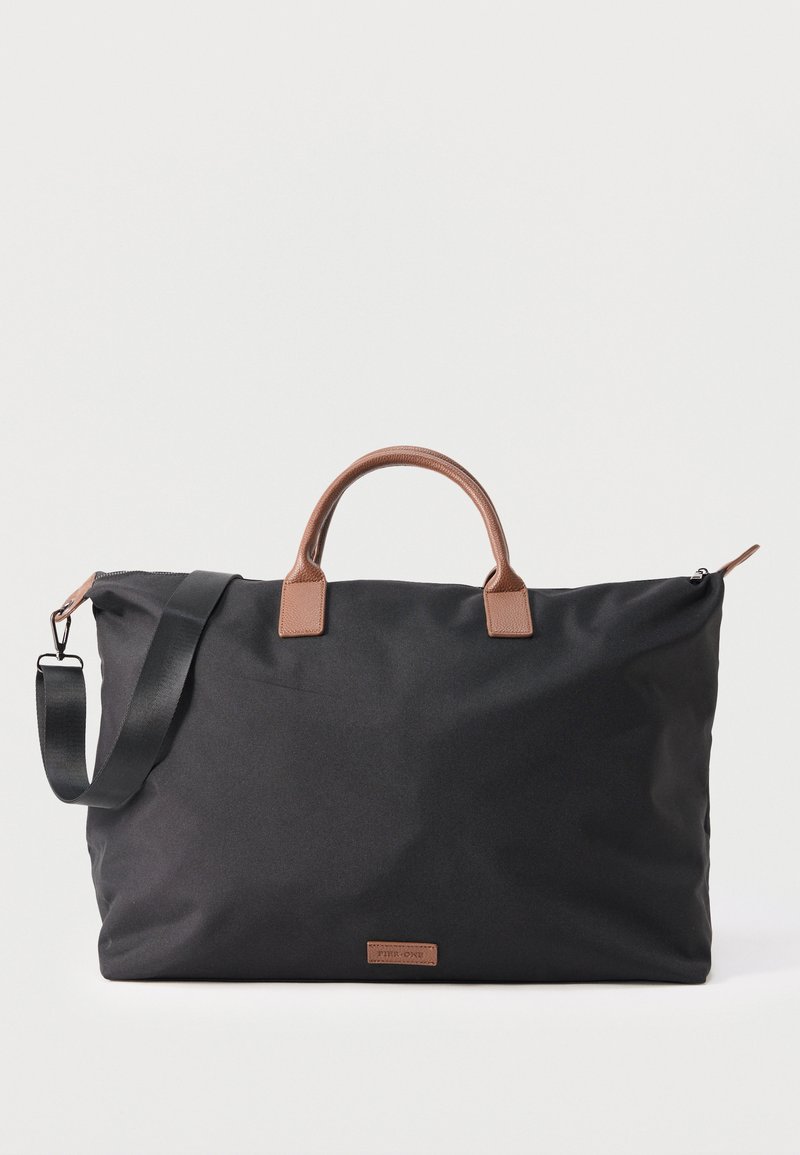 Μεγάλη μαύρη ταξιδιωτική τσάντα με καφέ δερμάτινες λαβές και αποσπώμενο μαύρο ιμάντα ώμου σε λευκό φόντο.