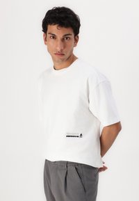White cotton t-shirt with a crew neckline, relaxed fit, and short sleeves. Features a black printed label on the lower left side.
