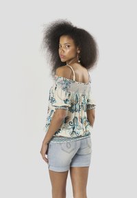 Top ligero con estampado floral y escote off-the-shoulder en color crema con detalles en azul y naranja; combinado con shorts de mezclilla clara, mostrando un ajuste relajado.