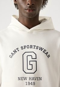 Sudadera blanca con cordón, que presenta un logo impreso en negro que dice "GANT SPORTSWEAR" y "NEW HAVEN 1949" en un estilo de fuente clásica.