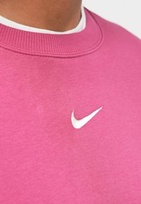Rozā džemperis ar ribotu apkakli; priekšpusē ir balts Nike logo. Mīksta auduma ar gludu tekstūru.