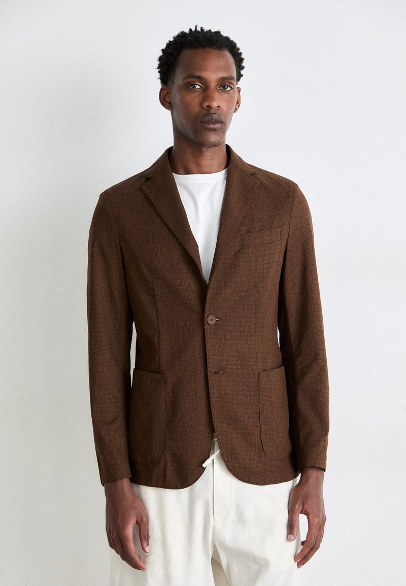Blazer marrone con texture, a un bottone, con rever a punte e due tasche frontali, indossato sopra una t-shirt bianca.