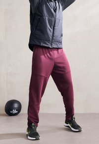 Bordeauxfarbene Sporthose aus strukturiertem Stoff, mit Seitennähten und elastischen Bündchen, kombiniert mit einer schwarzen Steppweste und dunklen Sneakers.