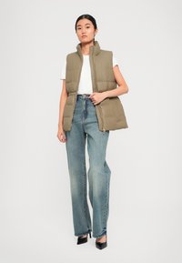 Gilet imbottito khaki con colletto alto, vita cinched, abbinato a una t-shirt bianca e jeans a vita alta di un blu chiaro. Scarpe nere con tacco.