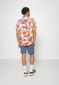Chemise à manches courtes beige clair avec un motif floral vif rouge et bleu. Assortie à un short bleu et des chaussettes blanches, se tenant sur un fond neutre.