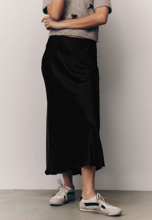 Isik kannab halli lühikeste varrukatega kampsunit, musta midi-seelikut ja valgeid tossusid ning seisab tavalise seina ees.