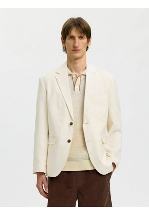 Mann trägt einen cremefarbenen Blazer über einem cremefarbenen Strick-Poloshirt und braunen Hosen, steht vor einem einfarbigen hellen Hintergrund mit einer Hand in der Tasche.