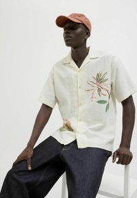 Camisa de manga corta, color amarillo pálido, con bordado floral en naranja y verde, combinada con pantalones de mezclilla azul oscuro. Modelo sentado en una silla.