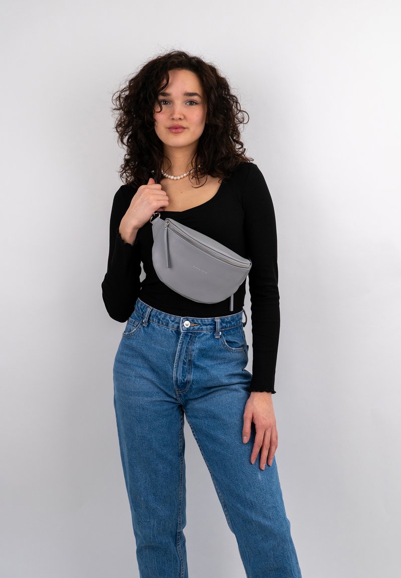 Borsa a fanny pack grigia e testurizzata indossata in vita, con chiusura a zip e dettaglio del logo, abbinata a una maglietta nera a maniche lunghe e jeans blu.
