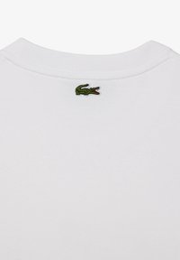 Col en tissu blanc avec un petit logo de crocodile vert brodé centré sous le col.