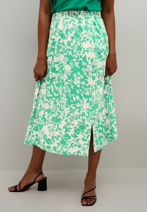 Bloemenrok met een groen-wit patroon, mid-lengte met een zijsplit, gemaakt van lichtgewicht stof. Ges 市ptoeren met zwarte sandalen met bandjes.