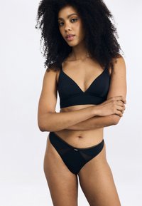 Melna bralette ar dziļu V-veida kakla izgriezumu un plecu siksnām, kombinēta ar atbilstošām melnām stringbiksēm ar mežģīņu akcentiem un siets detalizāciju.