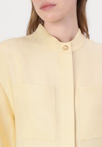 Ljusgul skjorta med knappslå och hög krage, två bröstfickor och en texturerad tyg. Har en mittknappstängning.