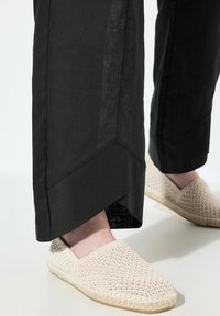 Svarta, vida byxor med en strukturerad yta har en distinkt vinklad fåll; ihop med beige vävda skorslips med ett mönstrat ovansida.