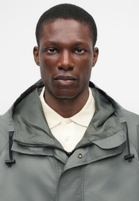 Πράσινο αδιάβροχο μπουφάν με φερμουάρ μπροστά, ρυθμιζόμενη κουκούλα και γιακά πάνω από ένα ανοιχτόχρωμο πουκάμισο. Λείος υφασμάτινος ιστός με ελάχιστες ραφές.