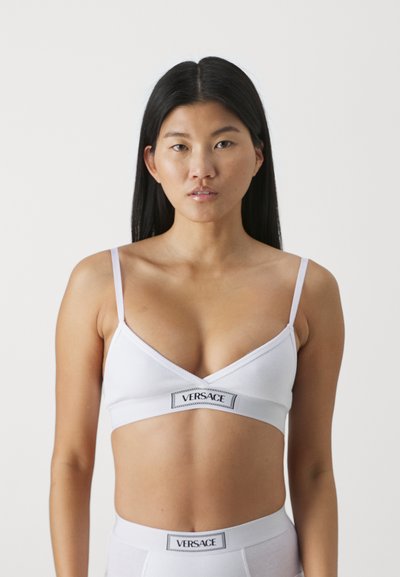 Weißes geripptes Bralette mit verstellbaren Trägern, das auf der Vorderseite ein schwarzes Versace-Logo trägt. Einfaches Design, weicher Textur und dreieckige Form.