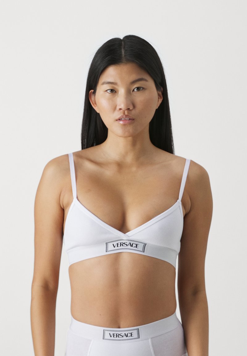 Hvit ribbestrikket bralette med justerbare stropper, med en svart Versace-logo foran. Enkel design, myk tekstur og trekantet form.