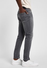 Hombre vestido con jeans ajustados grises y zapatillas blancas, con la mano descansando en el bolsillo trasero, de pie contra un fondo claro y liso.