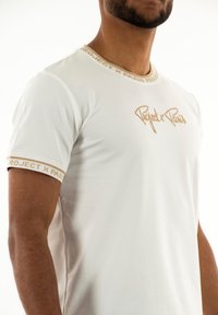 Camiseta blanca con cuello redondo, adornada con un ribete dorado y un logo. Tejido suave, mangas cortas y un diseño ajustado.
