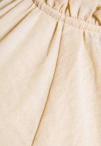 Ljust beige tyg med en mjuk textur, försedd med rynkad elastik i midjan och en subtil glans, arrangerad i flödande veck.