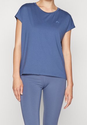 Femme portant un haut de sport bleu ajusté à manches courtes et des leggings assortis, debout devant un fond clair uni.