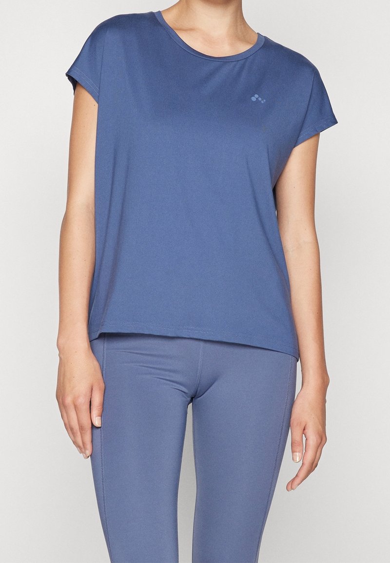 Femme portant un haut de sport bleu ajusté à manches courtes et des leggings assortis, debout devant un fond clair uni.