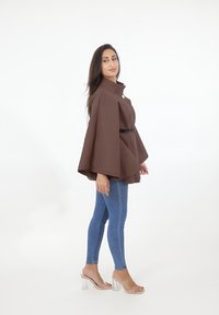 Blusa marrón de estilo capa con cuello alto, que presenta un botón plateado y un cinturón negro, combinada con jeans skinny azules y sandalias transparentes de tacón bloque.