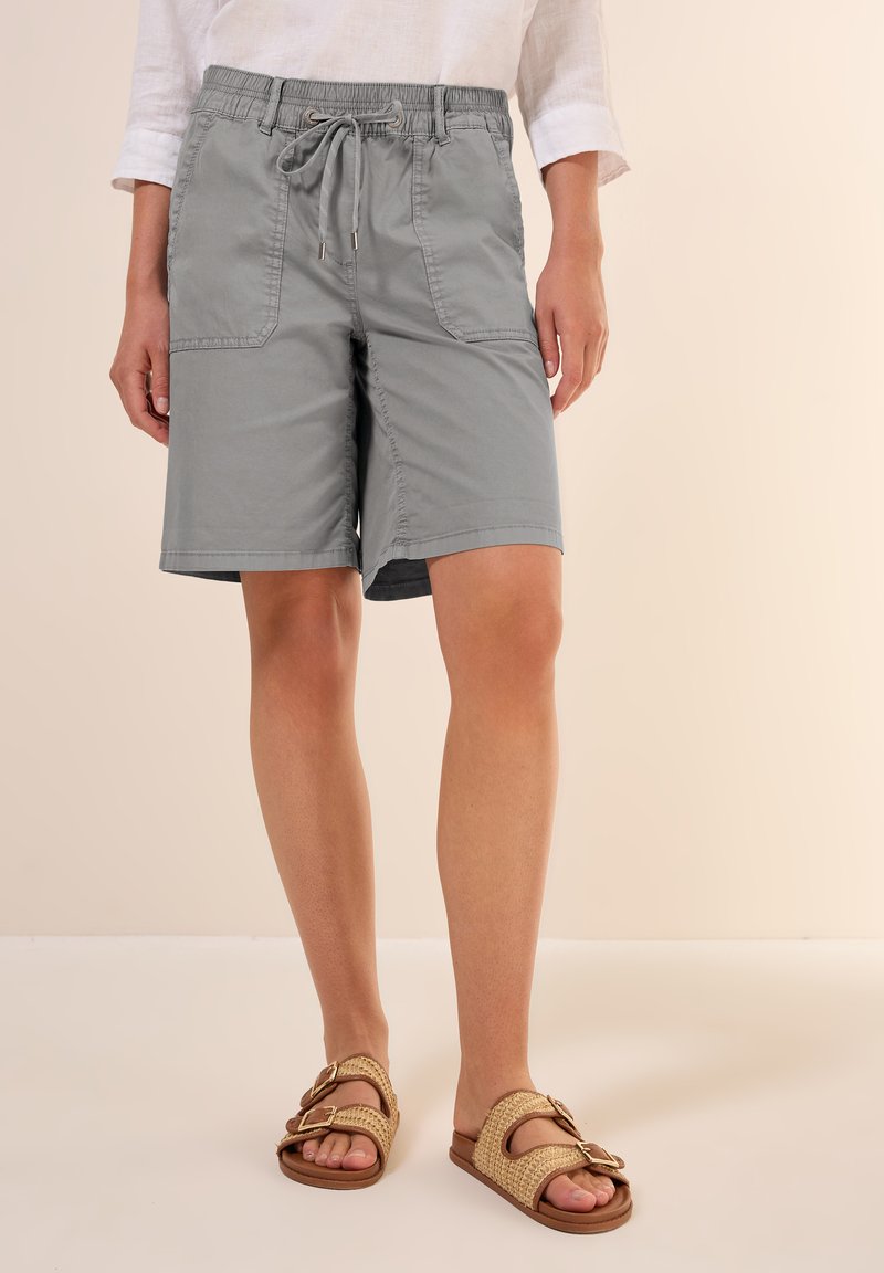 Shorts gris clair avec une ceinture élastique et un cordon de serrage, dotés de deux poches avant. Portés avec des sandales à enfiler tissées beiges.