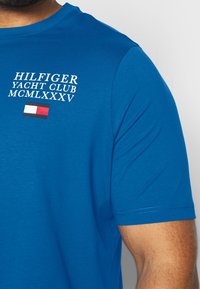 Modré tričko s krátkým rukávem, na kterém se nachází bílý text "HILFIGER YACHT CLUB MCMLXXXV" a malý červený, bílý a modrý emblém na přední straně.