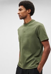 Olīvzaļš īsroku t-krekls no mīksta auduma ar diskrētu logo uz krūtīm un apaļu kakla izgriezumu. Teksturēts viendabīgs dizains.